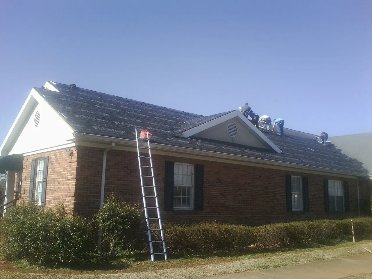 Crew tearing shingles off a brick ranch roof with a ladder set — Emergency Roof Repair work by our crew in Mannsville